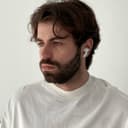 A man with wavy brown hair and a beard, wearing a white crewneck sweatshirt and white wireless earbuds, looks slightly to the left with a neutral expression. The background is plain and light-colored